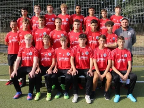 Das Foto zeigt die B1 Jugend des TSV Ronsdorf in der Saison 2022/2023 beim offiziellen Mannschaftsfoto. Die Spieler tragen ihre roten Trainingsshirts mit Sponsorenlogo und posieren gemeinsam mit ihren Trainern vor dem Tor.